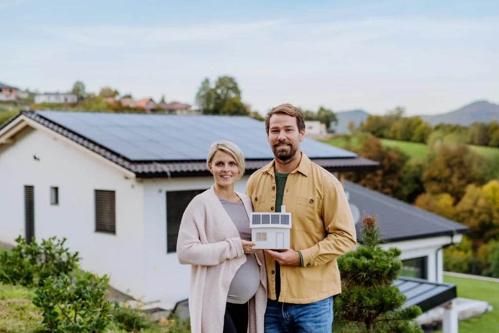 Couple tenant une maquette de maison devant une habitation équipée de panneaux solaires